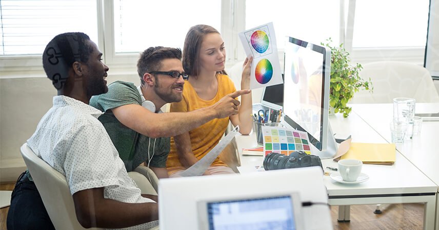 Drie mensen zitten rond een bureau en werken samen aan een project voor hun brandingbureau. Een man met een bril wijst naar een computerscherm met kleurenwielen, terwijl een vrouw een kleurenkaart omhoog houdt. Op het bureau liggen ook verschillende papieren, een camera en planten, die de essentie van creatieve groei vastleggen. Drie mensen zitten rond een bureau en werken samen aan een project voor hun brandingbureau. Een man met een bril wijst naar een computerscherm met kleurenwielen, terwijl een vrouw een kleurenkaart omhoog houdt. Op het bureau liggen ook verschillende papieren, een camera en planten, die de essentie van creatieve groei vastleggen.