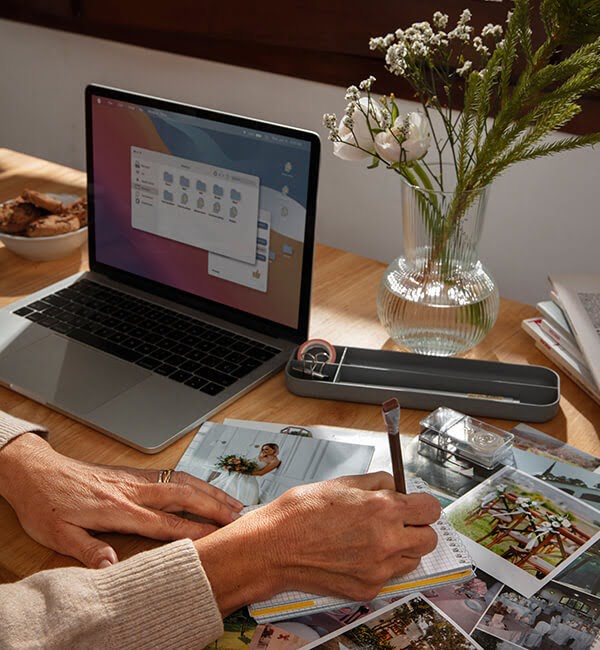 Een persoon werkt aan een plakboek aan een houten tafel met een laptop, foto's en een metalen kist met kunstbenodigdheden. Op tafel staat een vaas met bloemen en een schaal met koekjes. Zonlicht filtert door een raam en creëert een gezellige werkruimtesfeer die perfect is voor planning met hun brandingbureau.