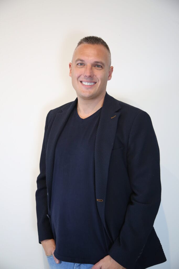 Lachende man in een donker jasje en t-shirt, staande tegen een effen witte achtergrond.