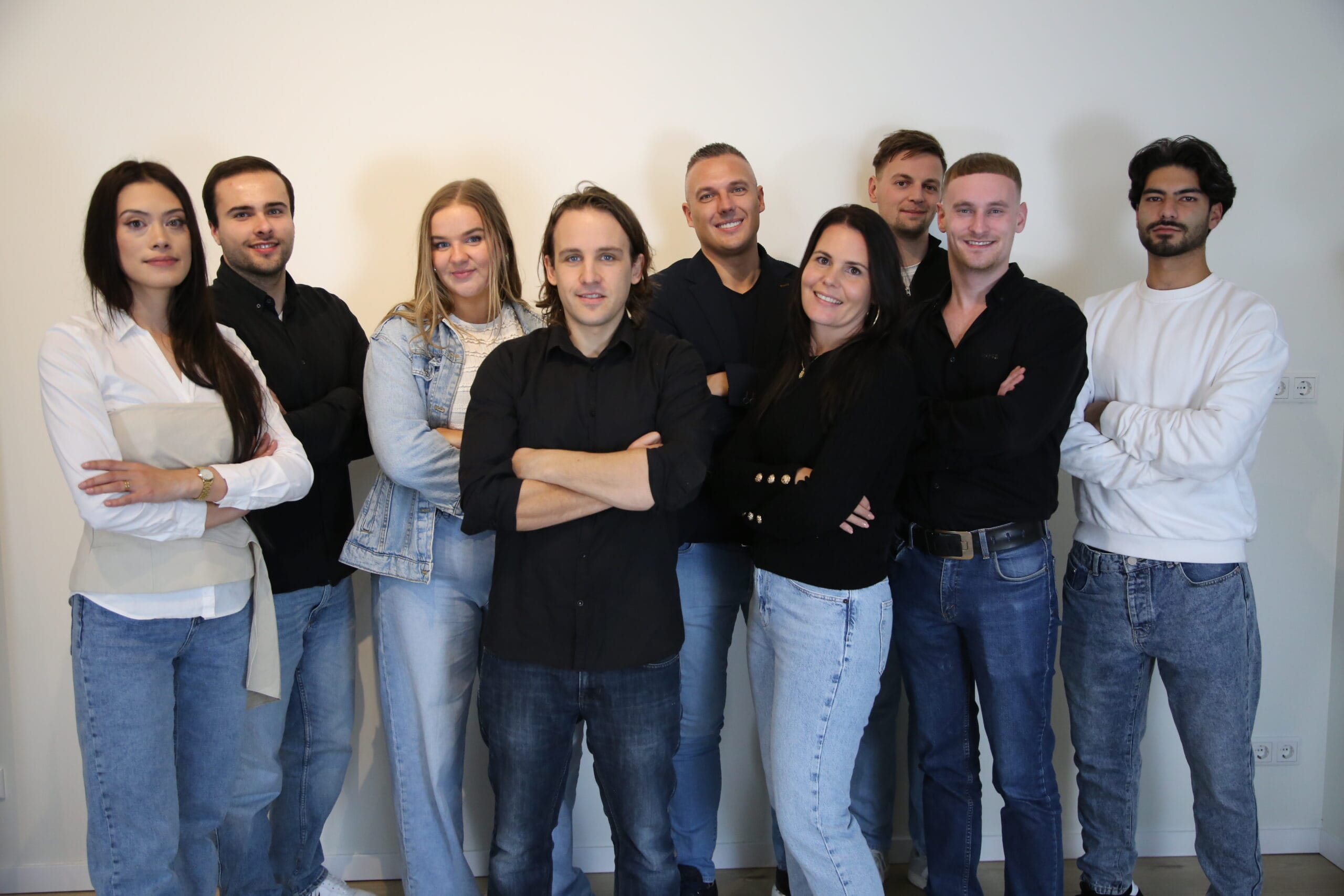 Een groep van negen mensen die samen staan, gekleed in casual outfits, sommigen in jeans en effen shirts. Ze poseren tegen een neutrale achtergrond, de meesten lachend, armen gekruist of ontspannen langs hun lichaam.