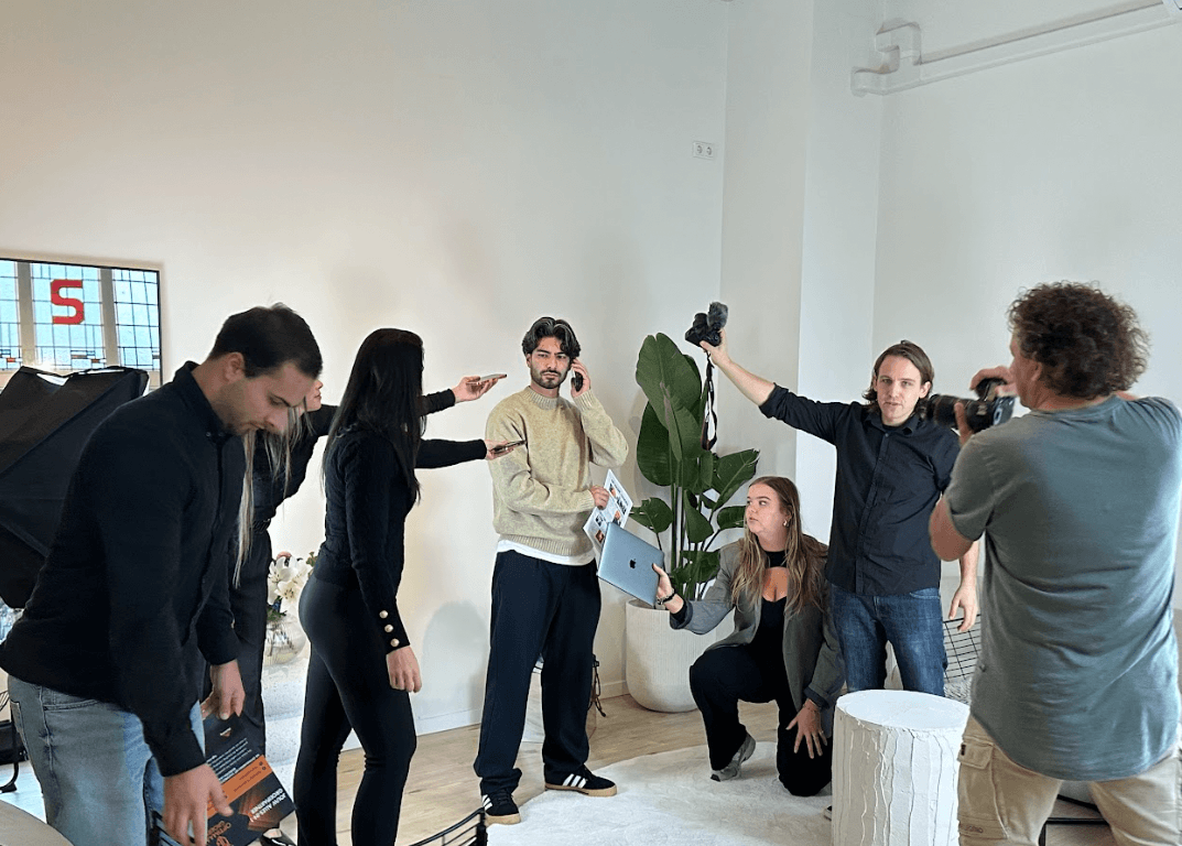 Een groep mensen is bezig met een fotoshoot in een goed verlichte kamer. De centrale persoon staat met een telefoon en een tijdschrift in zijn handen, terwijl anderen de belichting aanpassen en apparatuur om hem heen positioneren. Een fotograaf legt de scène vast.